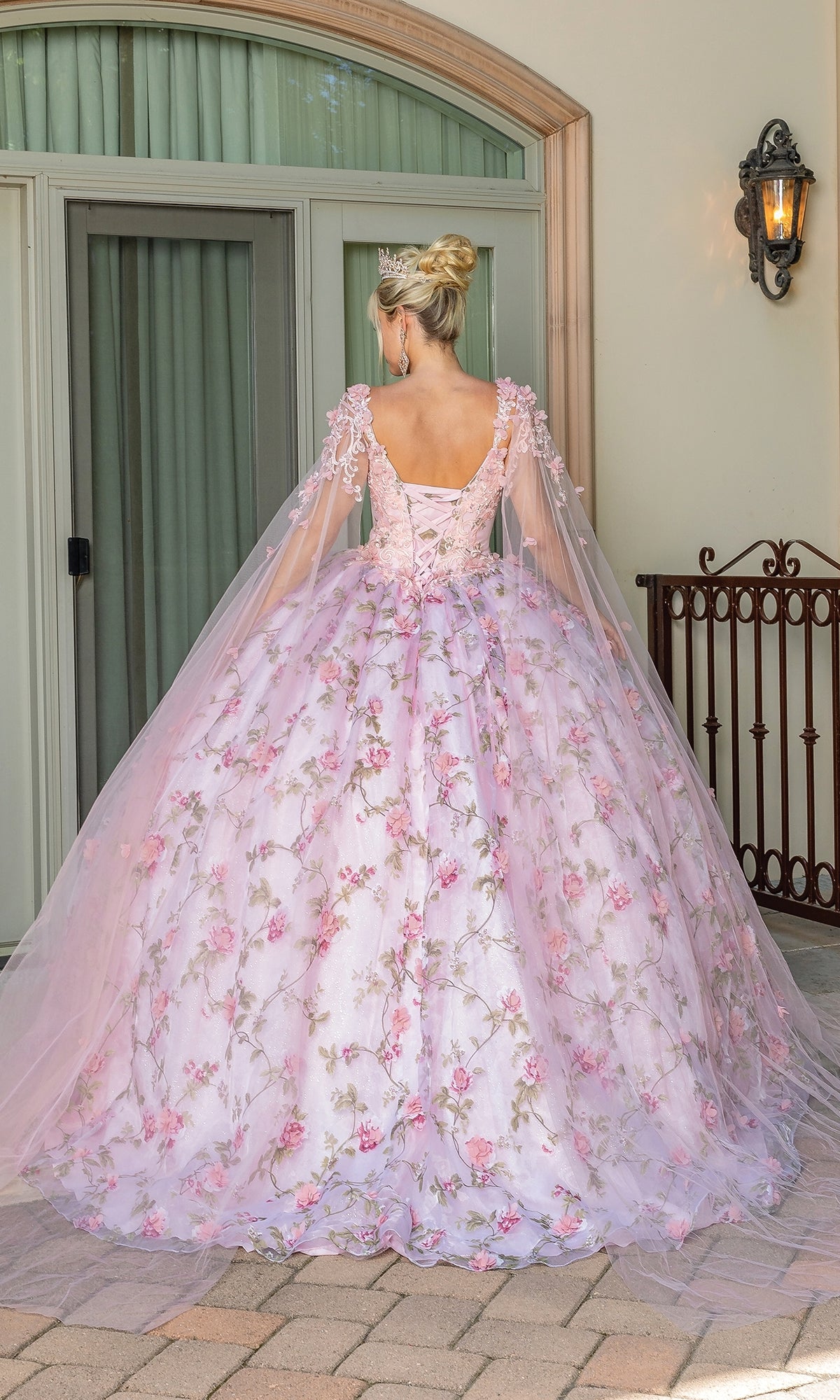 A woman stands outdoors facing away, wearing the Quinceanera Dress 1715 By Dancing Queen—a blush pink floral ball gown with lace-up back, tulle cape sleeves, and a tiara—in front of glass doors and a curved window.